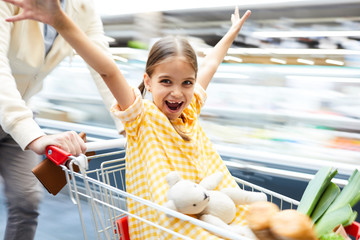 Jolly excited girl in checkered dress sitting in shopping cart and outstretching arms while having...