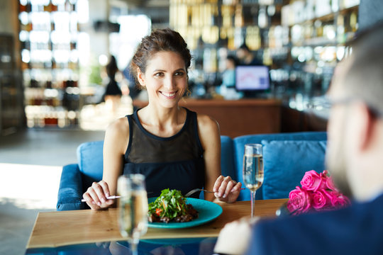 Smiling brunette girl eating fresh vegetable salad in restaurant and talking to her boyfriend during date