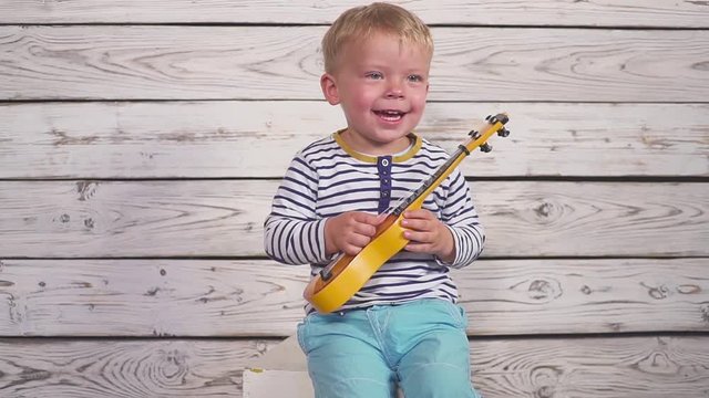 happy two year old boy plays his guitar or ukulele and sings songs, sitting in the wooden room.