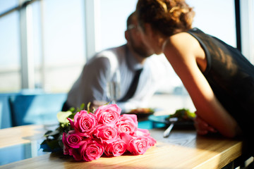 Bunch of pink roses on sunlit table and young amorous couple leaning over table for kiss - obrazy, fototapety, plakaty