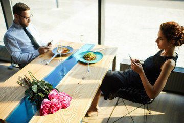 Contemporary young man and woman in elegant clothes sitting by served table in restaurant and using...