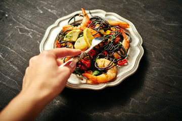 a woman's hand and delicious black spaghetti with various grilled shrimps and vegetables, on silver plate