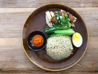 Roasted chicken breast with hainan rice and Bok choy and half egg with red hot sauce on brown plate on wooden table