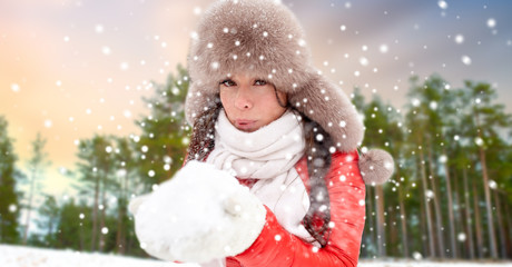 people, season and leisure concept - happy woman in fur hat blowing on snow in her hands over winter forest background