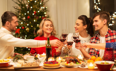 holidays and celebration concept - happy friends having christmas dinner at home, drinking red wine and clinking glasses
