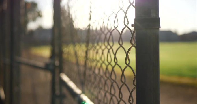 Close up of a chain link fence gate with locks on it at sunrise outside of a grass baseball field in a public park.