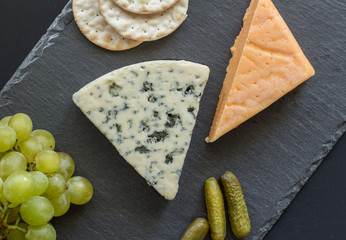 Cheese board with blue cheese, cheddar cheese, green grapes, crackers and pickled cucumbers on black slate - top view photograph