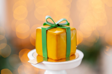Mini mousse pastry dessert with yellow glazed. garland lamps bokeh background. In the form of gift box, ribbons of chocolate. Modern european cake. French cuisine. Christmas theme