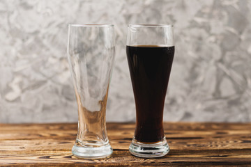 One empty glass next to full glass of soda water on wooden table on background of cement wall. Concept of leisure