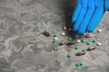 Human hand in blue rubber glove shows on scattered pills and capsules on gray cement background. Healthcare concept