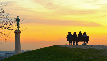silhouettes of people on the bench with Viktor column and statue,  cloudy colorful sunset in Belgarde, Serbia