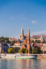 Two large Catholic churches on the Danube River in Budapest. The ship sails on the river. Many green plants on the Buda hill.