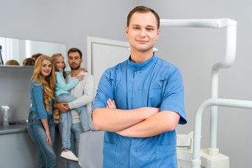 Fototapeta premium handsome young dentist with crossed arms looking at camera while young family standing on background together