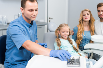 Obraz premium handsome young dentist with tools working with little child while her parents sitting near her