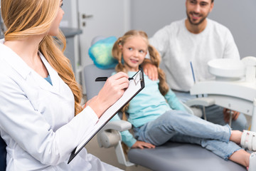 Fototapeta premium female dentist with clipboard talking to little child and her young father