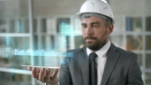 Medium shot of professional engineer in formal suit and hard hat holding futuristic gadget with spinning holographic projection of building plan and data