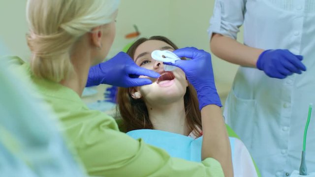 Dentist with assistant preparing girl for whitening teeth. Dentist installing rubber dam cheek retractor. Woman with open mouth sitting in dentist chair. Professional whitening procedure in clinic