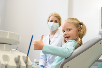 beautiful little child showing thumb up while sitting in chair at dentist office