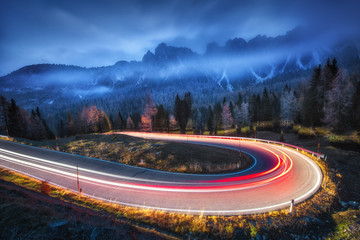 Phares de voiture flous sur route sinueuse dans les montagnes avec des nuages bas la nuit en automne. Paysage spectaculaire avec route goudronnée, sentiers lumineux, forêt brumeuse, rochers et ciel bleu. Voiture roulant sur la chaussée