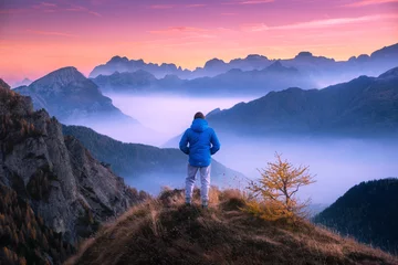 Fotobehang Chocoladebruin Sportieve man op de bergtop op zoek op bergdal met lage wolken bij kleurrijke zonsondergang in de herfst in de Dolomieten. Landschap met reiziger, mistige heuvels, bos in de herfst, geweldige lucht in de schemering in Alpen  © den-belitsky
