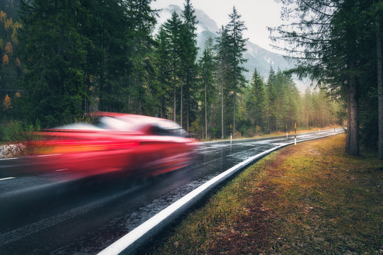 Blurred Red Car In Motion On The Road In Autumn Forest In Rain. Perfect Asphalt Mountain Road In Overcast Rainy Day. Roadway, Pine Trees In Italian Alps. Transportation. Highway In Foggy Woodland