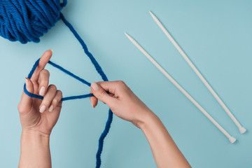 partial view of woman with blue yarn and white knitting needles knitting on blue backdrop