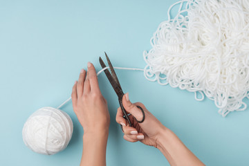 cropped shot of woman cutting white knitting thread with scissors on blue backdrop