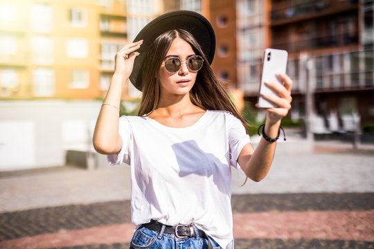 Outdoor street style image of young trendy woman making selfie on the street , wearing stylish hipster hat playing with her hairs.