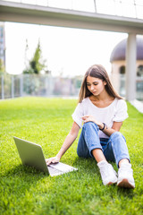 Young woman with laptop sitting on green grass and looking to a display outdoors