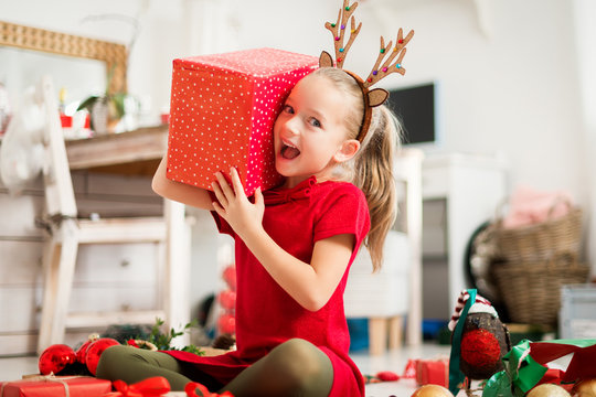 Cute super excited young girl opening large red christmas present while sitting on living room floor. Candid family christmas time lifestyle background.