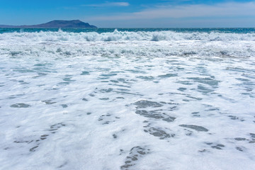 Beautiful turquoise waves with foam on a sandy beach, against the blue sky with clouds and a mountain on the horizon