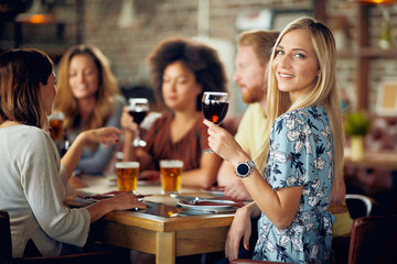 Woman looking at camera and holding glass of wine while sitting in restaurant. In background friends drinking and chatting.