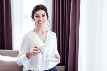 Tasty cappuccino. Joyful attractive woman holding a cup with coffee while looking at you