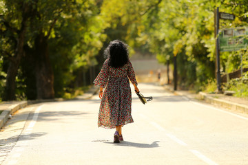 brunette girl with long hair in a fluttering long dress goes away on the road among the green trees