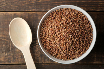 Dry buckwheat groats in a white bowl with a spoon on a wooden brown table. cereals. healthy food. porridge. top view - obrazy, fototapety, plakaty