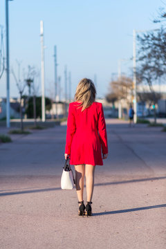 Rear image of a blonde woman wearing red jacket white handbag while walking in the city