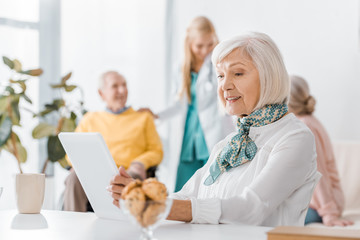 senior woman using digital tablet at nursing home with blurred people at background
