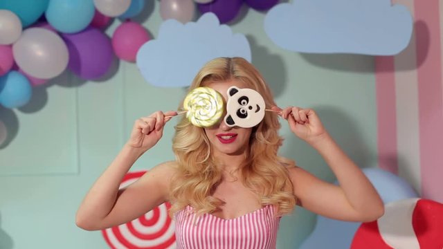A cheerful girl with big lollipops in her hands in a bright room with balloons and a large plastic ice cream, she covers her eyes with big lollipops. Barbie girl. Candy girl.