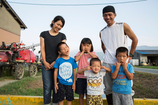 Portrait of smiling Japanese farmer, his wife and four children standing in their yard, children pulling faces at camera.