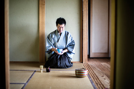 Japanese man wearing traditional kimono kneeling on tatami mat, holding tea bowl, during tea ceremony.