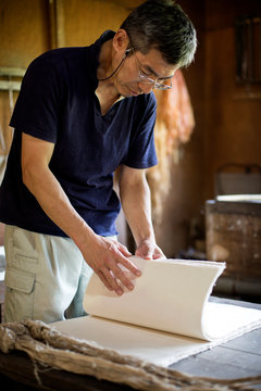 Japanese man standing in a workshop,  checking through sheets of traditional Washi paper.