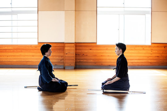 Female and male Japanese Kendo fighters kneeling opposite each other on wooden floor.