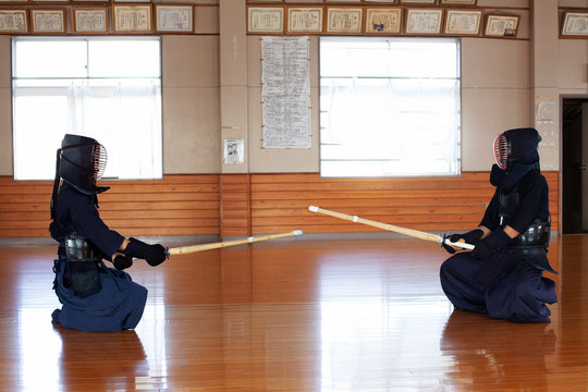 Two Japanese Kendo fighters sitting opposite each other on wooden floor, wearing Kendo masks and holding wood swords.