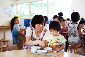 Female teacher sitting at table in a Japanese preschool, helping young boy to eat his lunch. 