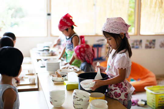 A girl and a boy wearing headscarves standing at a table in a Japanese preschool, serving lunch.