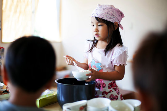 Girl wearing headscarf standing at a table in a Japanese preschool, serving lunch to children.