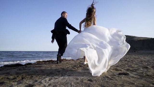 Following young couple in bridal dress and suit running on sandy beach, beautiful white dress waving on the wind
