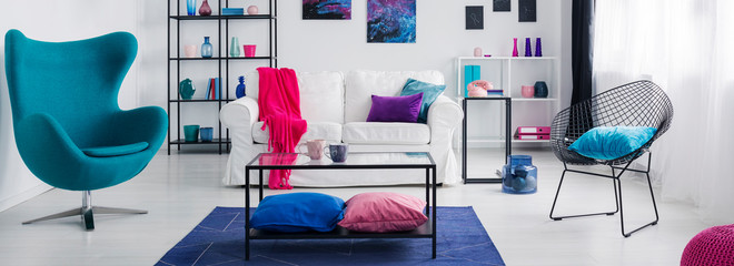 Table on blue carpet in white living room interior with armchair and pink blanket on sofa. Real photo