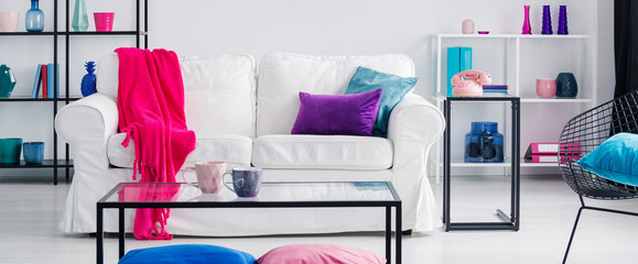 Pink blanket on white couch in flat interior with table and blue cushion on armchair. Real photo