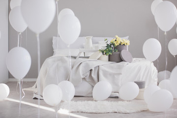 Flowers on bed with blanket and pillows in grey bedroom interior with white balloons. Real photo